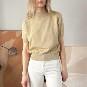 Könnte beinhalten: Ein hellbeiger Rollkragenpullover mit kurzen Ärmeln. Der Pullover hat ein dezentes, gesprenkeltes Muster und einen gerippten Saum.