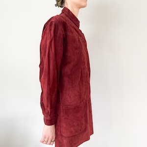 May include: A burgundy suede jacket with a button-up front, two front pockets, and long sleeves. The jacket has a high collar and a slightly worn appearance, suggesting a vintage style.