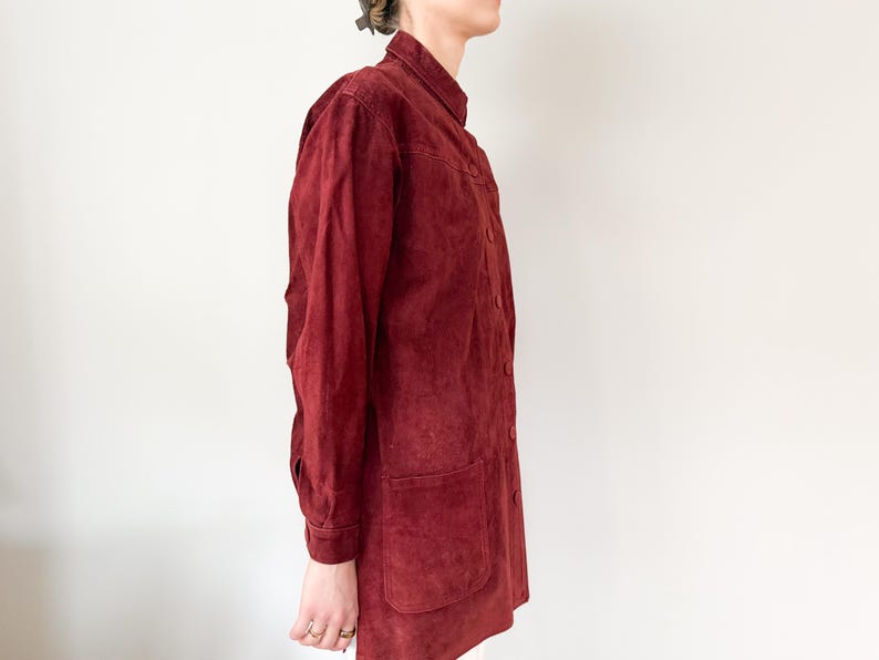 May include: A burgundy suede jacket with a button-up front, two front pockets, and long sleeves. The jacket has a high collar and a slightly worn appearance, suggesting a vintage style.