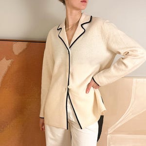 May include: Cream-colored blazer with black trim detailing. The blazer features a single-button closure and a classic lapel design. The sleeves are long, and the overall style is tailored. The blazer is paired with white pants.