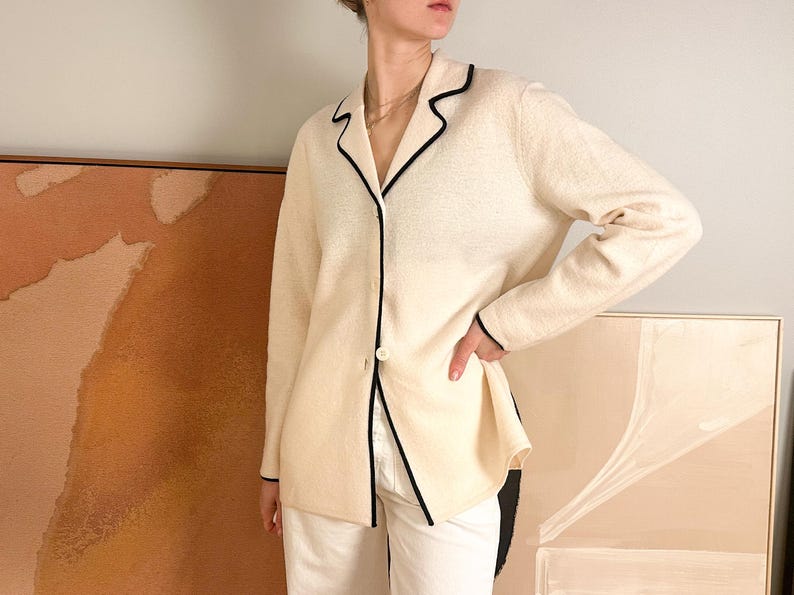 May include: Cream-colored blazer with black trim detailing. The blazer features a single-button closure and a classic lapel design. The sleeves are long, and the overall style is tailored. The blazer is paired with white pants.