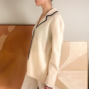 May include: Cream-colored blazer with black trim. The jacket has a classic collar and long sleeves. The blazer is paired with white pants. The background includes abstract art pieces in neutral tones.