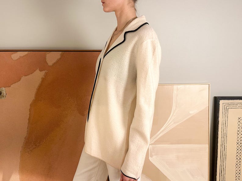 May include: Cream-colored blazer with black trim. The jacket has a classic collar and long sleeves. The blazer is paired with white pants. The background includes abstract art pieces in neutral tones.