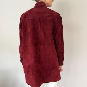 May include: A burgundy suede shirt with long sleeves and buttoned cuffs. The shirt has a collared neckline and a button-down front. The back of the shirt is visible, showing the texture of the suede fabric.