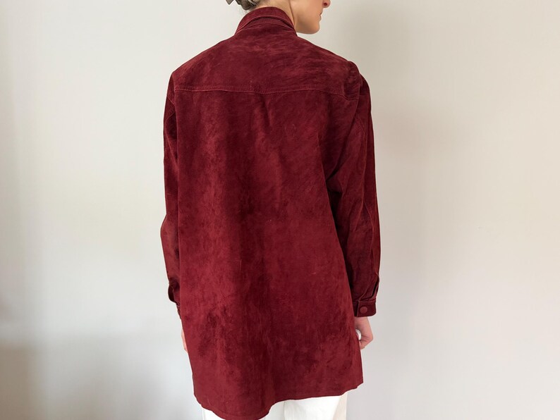 May include: A burgundy suede shirt with long sleeves and buttoned cuffs. The shirt has a collared neckline and a button-down front. The back of the shirt is visible, showing the texture of the suede fabric.
