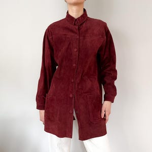 May include: A burgundy suede shirt-jacket with a button-down front, long sleeves, and two front pockets. The shirt-jacket has a collared neckline and is worn over white pants. The garment is a medium length.