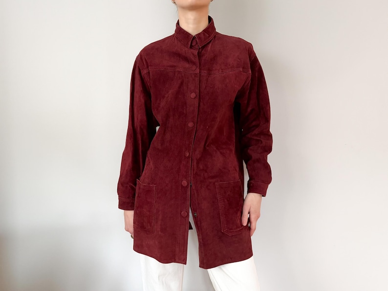 May include: A burgundy suede shirt-jacket with a button-down front, long sleeves, and two front pockets. The shirt-jacket has a collared neckline and is worn over white pants. The garment is a medium length.