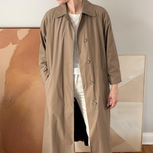 Puede incluir: Un abrigo largo tipo gabardina de color beige con cierre de botones y bolsillos laterales. El abrigo llega a la rodilla y tiene un cuello clásico. El modelo lleva debajo una camisa y pantalones blancos. El abrigo es de color neutro.