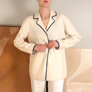 May include: Cream-colored blazer with black trim. The blazer has a classic lapel collar and a single-button closure. The sleeves are long, and the trim outlines the collar, lapels, and cuffs. The blazer is worn over white pants.