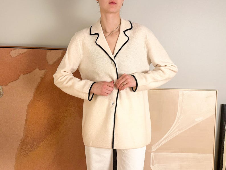May include: Cream-colored blazer with black trim. The blazer has a classic lapel collar and a single-button closure. The sleeves are long, and the trim outlines the collar, lapels, and cuffs. The blazer is worn over white pants.