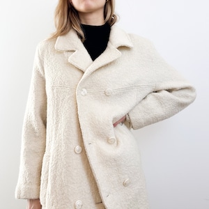 May include: A white double-breasted coat with a textured, fuzzy fabric. The coat has four large buttons and a classic collar.