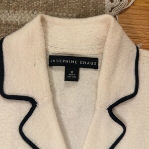May include: Close-up of a cream-colored jacket with a black trim around the collar and lapels. The label reads "JOSEPHINE CHAUS" and includes a size "M" tag. The jacket is made in Hong Kong.