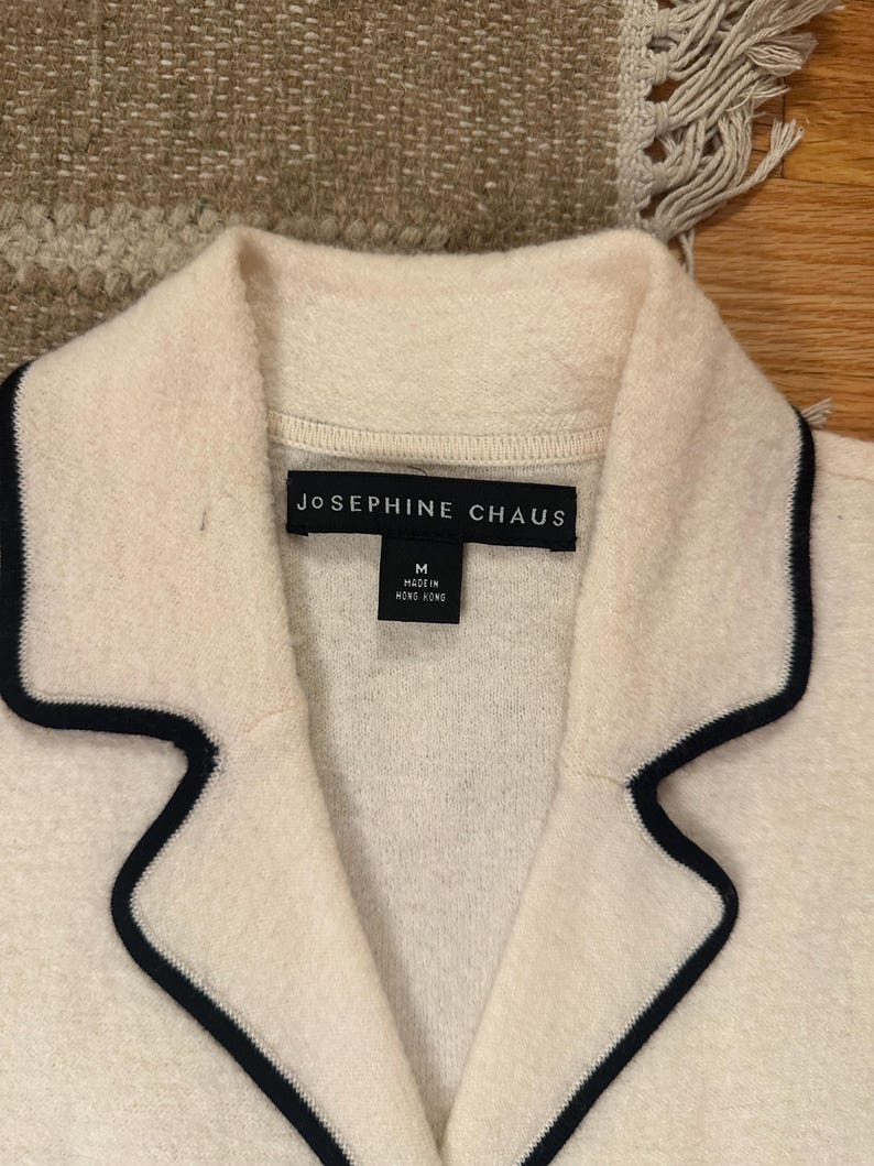 May include: Close-up of a cream-colored jacket with a black trim around the collar and lapels. The label reads "JOSEPHINE CHAUS" and includes a size "M" tag. The jacket is made in Hong Kong.