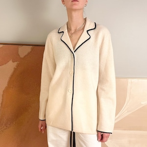 May include: Cream-colored button-down jacket with black trim. The jacket has a classic collar and long sleeves. The black trim outlines the collar, front, and cuffs. The jacket is worn over white pants.