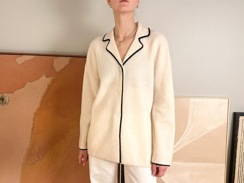 May include: Cream-colored button-down jacket with black trim. The jacket has a classic collar and long sleeves. The black trim outlines the collar, front, and cuffs. The jacket is worn over white pants.