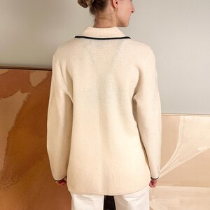 May include: Cream-colored sweater with a collared neckline and black trim. The long-sleeved sweater has a textured appearance and is worn by a person. The background includes artwork and a neutral wall.