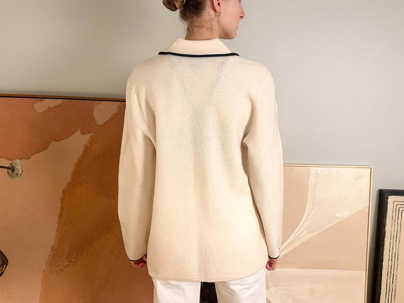 May include: Cream-colored sweater with a collared neckline and black trim. The long-sleeved sweater has a textured appearance and is worn by a person. The background includes artwork and a neutral wall.