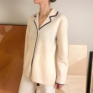 May include: Cream-colored pajama top with black trim. The long-sleeved top has a button-down front and a classic collar. The black trim outlines the collar, cuffs, and front closure. The top is paired with white pants.