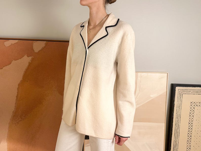May include: Cream-colored pajama top with black trim. The long-sleeved top has a button-down front and a classic collar. The black trim outlines the collar, cuffs, and front closure. The top is paired with white pants.
