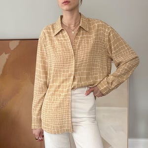Op de afbeelding: Een beige overhemd met knoopsluiting en een rasterpatroon. Het shirt met lange mouwen heeft een kraag en wordt gecombineerd met een witte broek. Het patroon van het shirt lijkt op een reptielenhuid. Het model draagt een ketting.