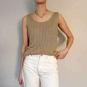 May include: A sleeveless, gold-colored knit top with a textured pattern. The top has a scoop neckline and is paired with white jeans. The overall style is casual and simple.