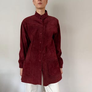 May include: A burgundy suede shirt with long sleeves, a collared neckline, and button-down closure. The shirt features two front pockets and is paired with white pants. The overall style is vintage.