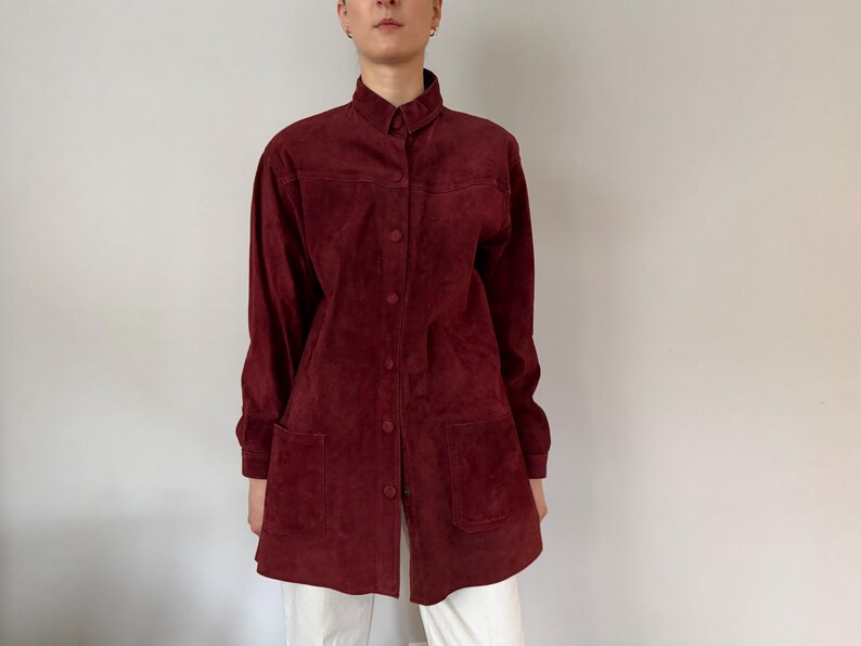 May include: A burgundy suede shirt with long sleeves, a collared neckline, and button-down closure. The shirt features two front pockets and is paired with white pants. The overall style is vintage.