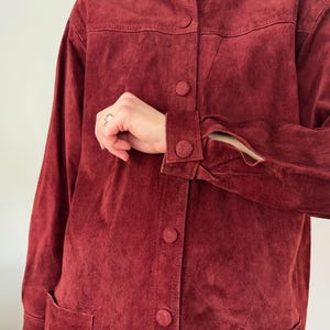 May include: A burgundy suede jacket with a button-up front and long sleeves. The jacket has a classic design with a collar and two front pockets. The cuffs have button closures. The jacket is a rich, solid color.