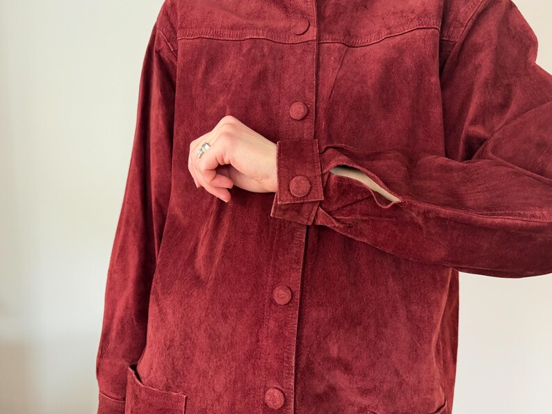 May include: A burgundy suede jacket with a button-up front and long sleeves. The jacket has a classic design with a collar and two front pockets. The cuffs have button closures. The jacket is a rich, solid color.