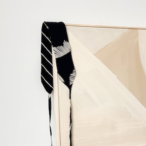 May include: A black and white patterned fabric strap hangs from a wooden frame. The strap has a striped pattern and a white leaf design. The frame is attached to a canvas with a beige background and abstract shapes painted in white.
