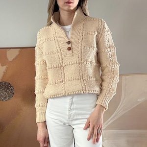 Op de afbeelding: Een crèmekleurige gebreide trui met een mock neck en een knoopsluiting. De trui heeft een textuurpatroon en is gecropped.