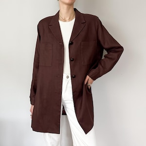 Puede incluir: Un blazer largo de color marrón oscuro con cuello de solapa clásico y dos bolsillos delanteros. El blazer tiene tres botones marrones oscuros y se usa sobre una camiseta blanca y pantalones blancos. El blazer es de estilo largo y entallado.