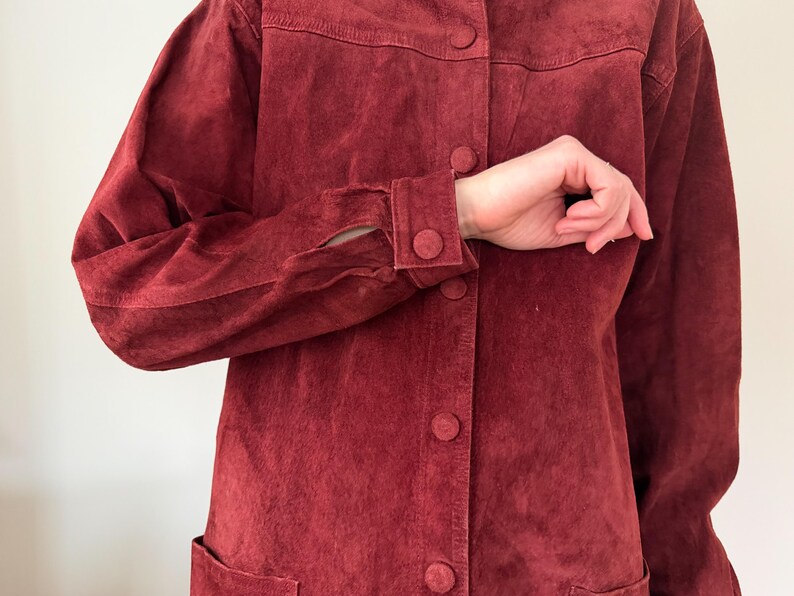 May include: A burgundy suede jacket with a button-up front, two front pockets, and buttoned cuffs. The jacket has a classic design and a soft, textured appearance. The sleeves are long and the jacket is a solid color.