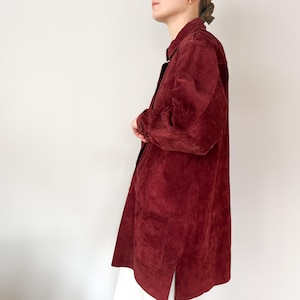 May include: A long, burgundy suede jacket with a collared neckline and two front pockets. The jacket has long sleeves and a button-down closure. The garment is knee-length and has a split at the bottom hem. The jacket is a solid color.