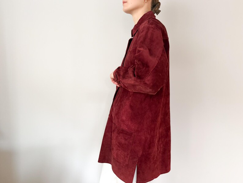 May include: A long, burgundy suede jacket with a collared neckline and two front pockets. The jacket has long sleeves and a button-down closure. The garment is knee-length and has a split at the bottom hem. The jacket is a solid color.
