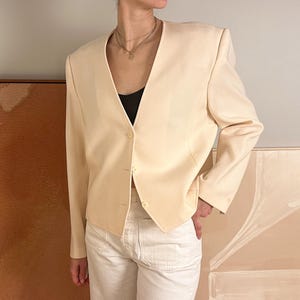 Op de afbeelding: Een crèmekleurige blazer met een V-hals en drie knopen. De blazer heeft gestructureerde schouders en lange mouwen. De persoon draagt een witte broek en een zwarte top. De achtergrond bevat abstracte kunst.