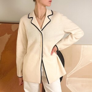 May include: Cream-colored blazer with black trim. The long-sleeved jacket has a notched collar and three buttons. The blazer is paired with white pants.