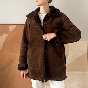 May include: A brown suede coat with a faux fur lining and trim. The coat has a button-up front, a collar, and long sleeves. The coat is knee-length and appears to be vintage. The coat is worn over white pants.