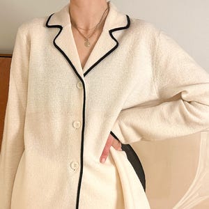 May include: Cream-colored button-down jacket with black trim along the collar, lapels, and pockets. The jacket has a textured fabric and a classic, tailored design. The buttons are a matching cream color. The jacket is part of a clothing collection.