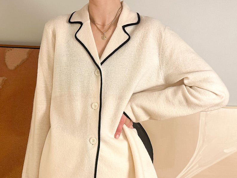 May include: Cream-colored button-down jacket with black trim along the collar, lapels, and pockets. The jacket has a textured fabric and a classic, tailored design. The buttons are a matching cream color. The jacket is part of a clothing collection.