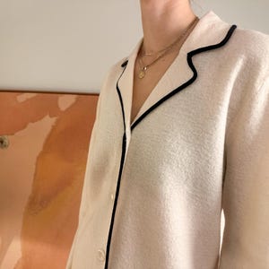 May include: Cream-colored jacket with black trim around the collar and lapels. The jacket has a button-down front and a textured fabric. A gold necklace is visible.