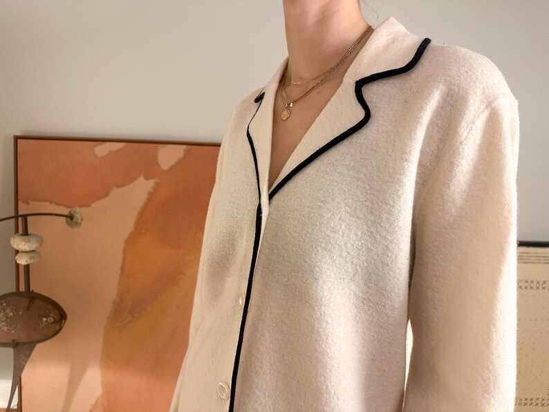 May include: Cream-colored jacket with black trim around the collar and lapels. The jacket has a button-down front and a textured fabric. A gold necklace is visible.