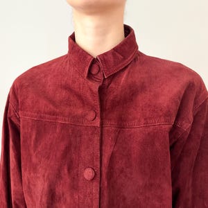May include: A burgundy suede jacket with a button-up front and a classic collar. The jacket has three visible buttons and a textured surface. The garment is a vintage piece.