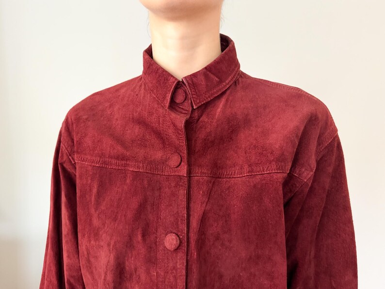 May include: A burgundy suede jacket with a button-up front and a classic collar. The jacket has three visible buttons and a textured surface. The garment is a vintage piece.
