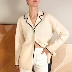 May include: Cream-colored button-down cardigan with black trim. The long-sleeved sweater has a V-neck and is paired with white pants. The cardigan's black trim outlines the collar, cuffs, and front edges, creating a classic look.
