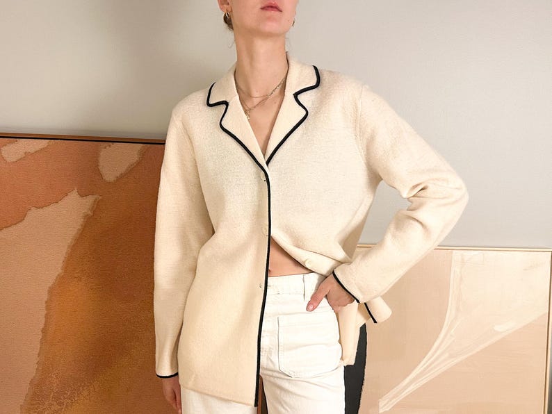 May include: Cream-colored button-down cardigan with black trim. The long-sleeved sweater has a V-neck and is paired with white pants. The cardigan's black trim outlines the collar, cuffs, and front edges, creating a classic look.
