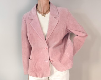 Puede incluir: Un blazer de ante rosa claro con solapa de muesca, mangas largas y cierre frontal de dos botones. La chaqueta incluye dos bolsillos delanteros inferiores y se lleva sobre una parte superior blanca acanalada con un collar de cadena dorada.