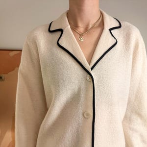 May include: Cream-colored jacket with black trim around the collar and lapels. The jacket has a button-down front and is paired with a gold necklace. The jacket is made of a textured fabric.
