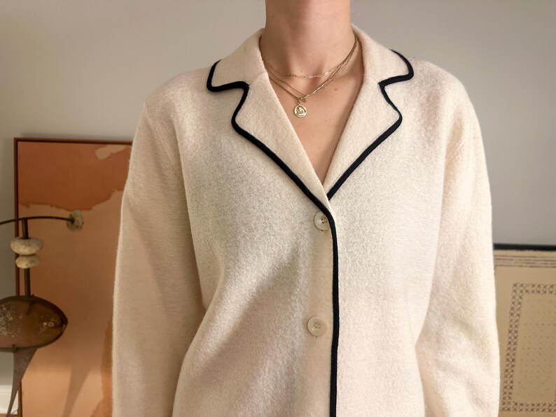 May include: Cream-colored jacket with black trim around the collar and lapels. The jacket has a button-down front and is paired with a gold necklace. The jacket is made of a textured fabric.