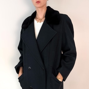 May include: A black double-breasted coat featuring a wide, plush black faux fur collar and long sleeves. The coat has two visible black buttons on the front and two side pockets. A white ribbed shirt is visible underneath the coat's open neckline.